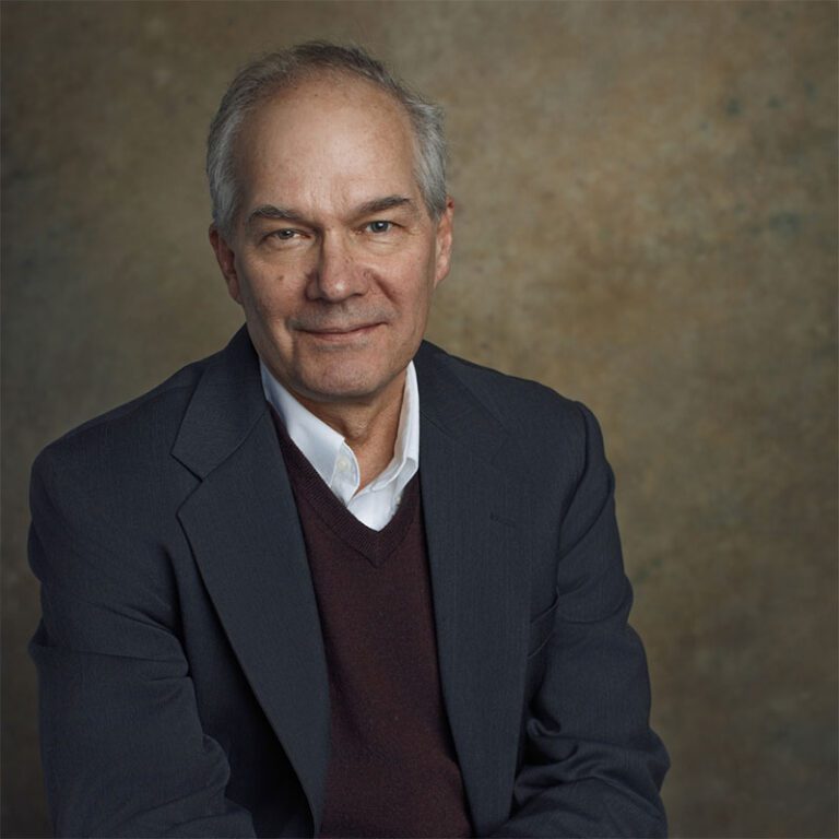Professional studio portrait of a man seated against a neutral mottled background, wearing a dark blazer over a dark sweater and a white collared shirt, facing the camera.